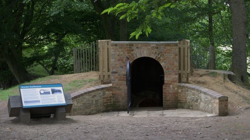 A brick fronted structure with an arched entranceway. A black metal gate is open. There is a sign to the left of the structure and trees in the background.